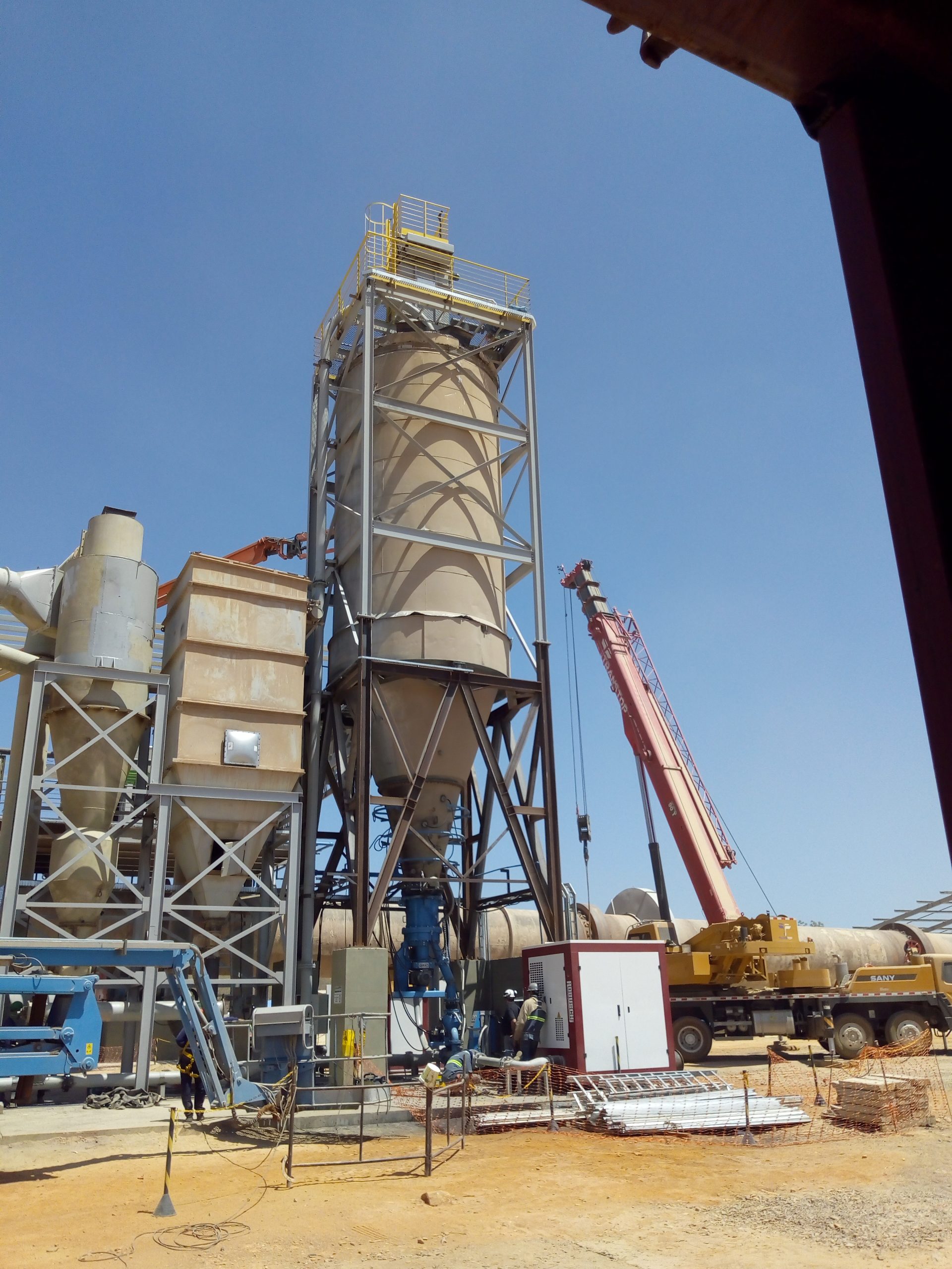 Balança industrial em um campo de chão de terra batida, sob um céu azul da roça. A balança parece um tanque, deve ter uns 3 metros de altura ou mais.
